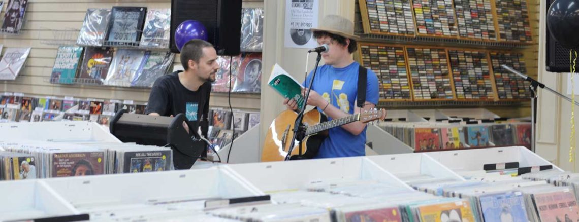 Image contains a photo of two people talking with each other in the background of a record store. One guy has an acoustic guitar and is standing behind a microphone while opening a notebook.