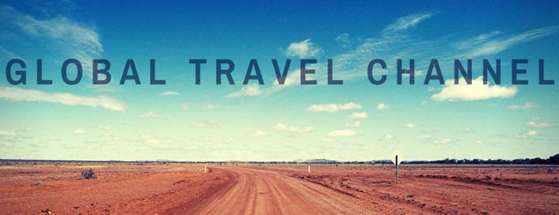 Image contains a photo of a long dirt road heading to the horizon. The sky above is mostly clear and blue with a few small white clouds. In the foreground, there is black text that says "Global Travel Channel".