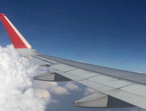 Image contains a photo of the wing of a plane with a blue sky above it and clouds below.