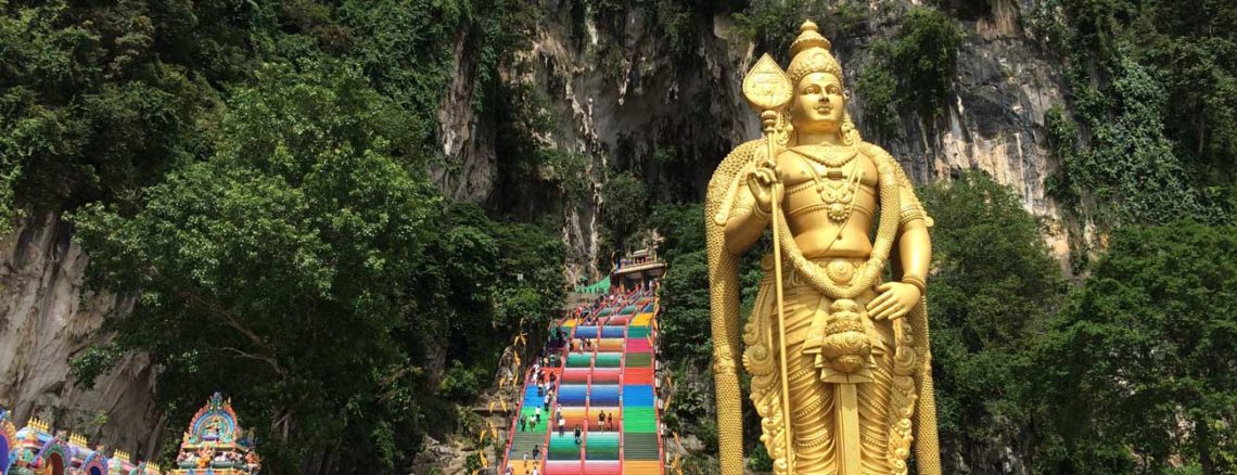 Image contains a photo of a large set of stairs ascending into a cave. The stairs are painted in bright colours and they're surrounded by jungle vegetation. In the foreground, to the right of the stirs, there is a large golden statue.