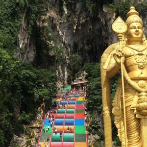 Image contains a photo of a large set of stairs ascending into a cave. The stairs are painted in bright colours and they're surrounded by jungle vegetation. In the foreground, to the right of the stirs, there is a large golden statue.