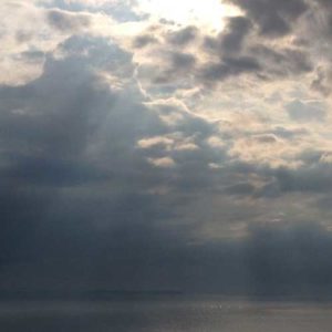 Image contains a photo of a large body of salt water extending beyond the horizon. Above the horizon line, there are clouds and a variety of sun beams breaking through them down to the water below.