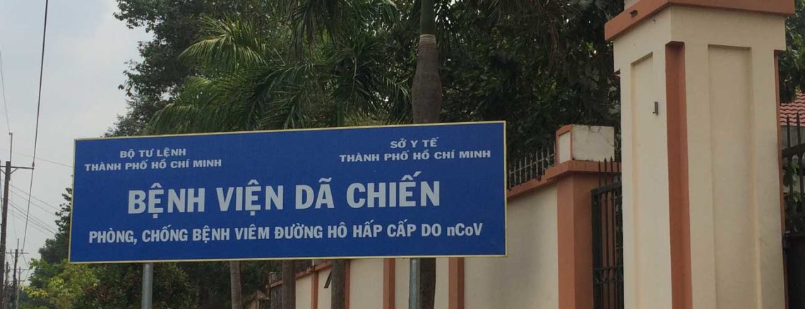 Image contains a photo of a large blue sign with white Vietnamese text. The sign is located just outside of a large wall and there are trees in the background.