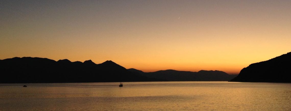 Image contains a photo of a sunset over a large body of water. On the horizon, hills can be seen stretching across most of the frame. The sky is a bright orange near the horizon, and it eventually turns a dark blue near the top of the frame.