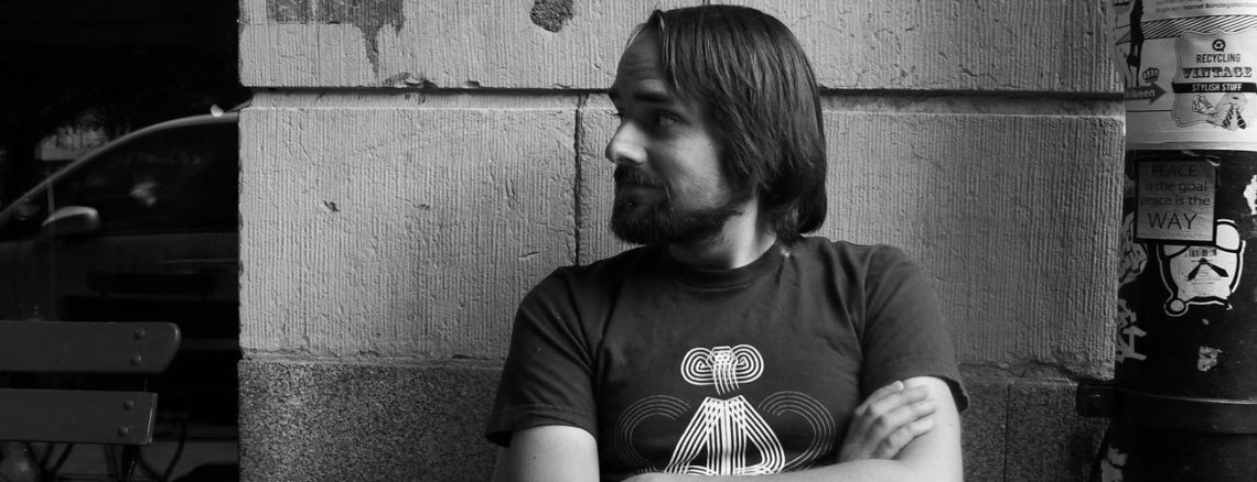 Image contains a black and white photo of a male looking sideways to the left of the camera. His arms are crossed and he's wearing a t-shirt. In the background, there is a large brick wall and a telephone pole to the right of the man. There are various posters on the pole.