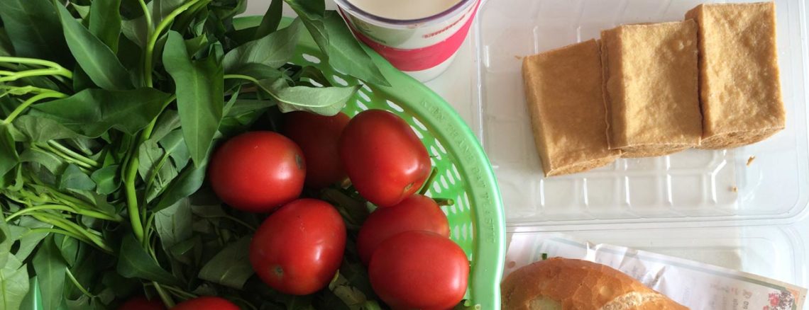 Image contains a photo of a variety of food on a white tabletop. On the left-hand side, in a green plastic basket, there are bright red tomatoes and a bunch of morning glory. To the right of the basket, there is a plastic container of small blocks of fried tofu, and just below that there is a banh mi sandwich.