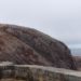 Image contains a photo of Signal Hill in St. John’s, Newfoundland and Labrador. In the foreground, an old cannon can be seen sticking out from the left, and there is a shot brick wall just in front of it. In the background, there is a hill with a small tower on top of it. Beyond that, there is a grey sky and the ocean.