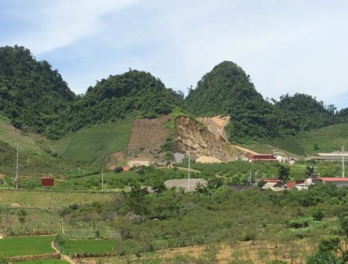 Image contains a photo of rolling hills and a green valley. In the foreground the peaks of several buildings with red tiled roofs can be seen.