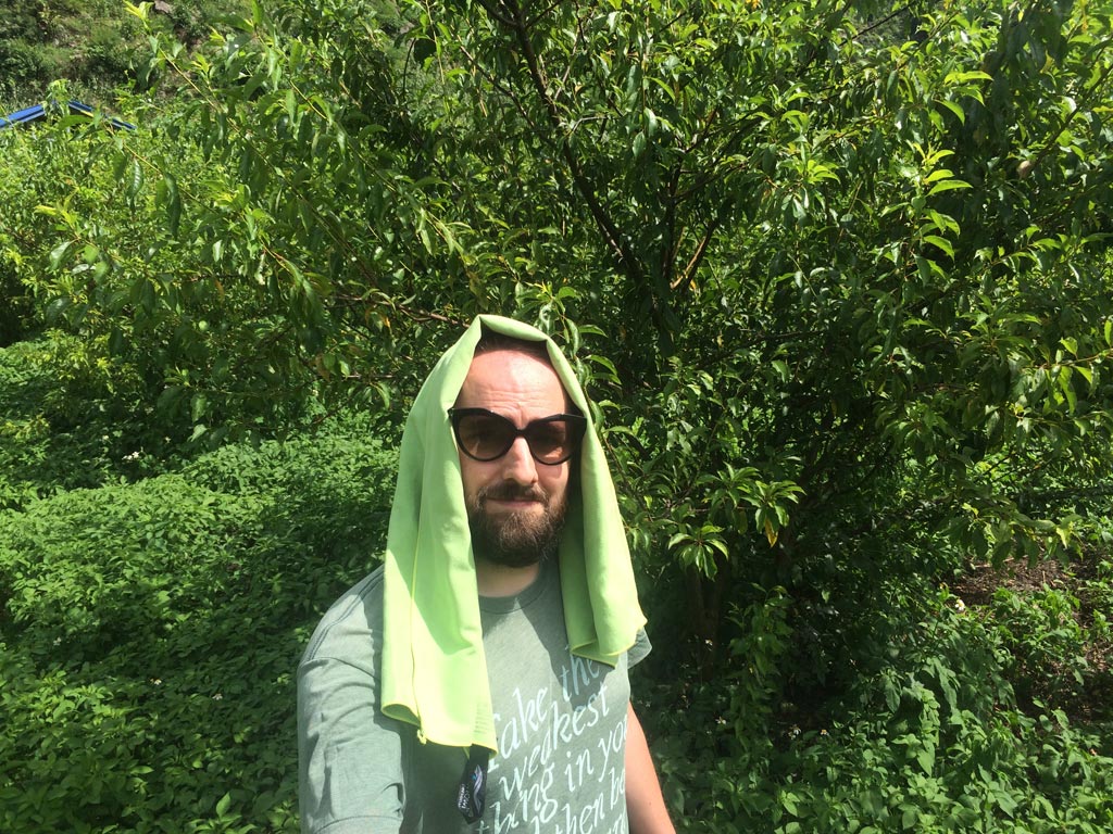 Image contains a photo of a bearded man wearing sunglasses and a bright green towel on his head. The man is surrounded by green leafy trees and plants.