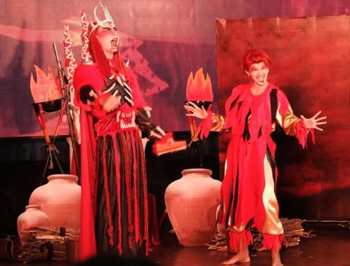 Image contains a photo of a theatrical production involving two characters in a setting similar to Hell. the male character is taller and wears a crown and armour, as well as a cape. The female character is striking a comedic pose with a smile on her face. Much of the photo is in red tones.