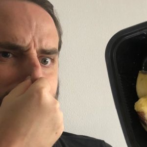 Image contains a photo of a bearded white man pinching his nose and looking unhappy. To the right of the man, there is a black plastic tray of durian, which is a yellowish fruit shaped in large pieces.