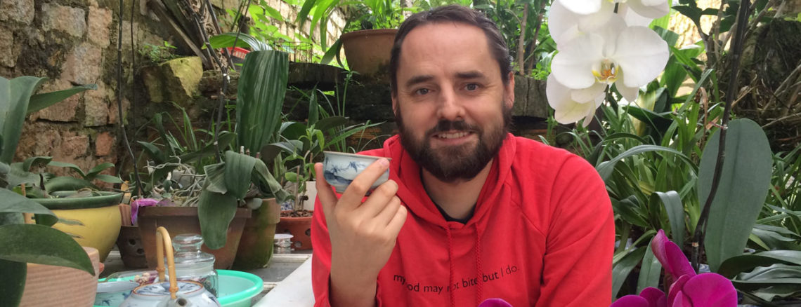 Image contains a photo of a bearded white man in a red hoodie sitting and holding a small tea cup. The man is smiling at the camera and there are plants surrounding him. To the left of he man, there is a white door being used as a table with a numbed of potted plants resting on it.