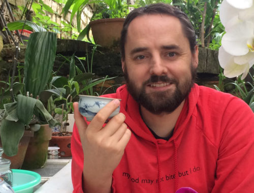 Image contains a photo of a bearded white man in a red hoodie sitting and holding a small tea cup. The man is smiling at the camera and there are plants surrounding him. To the left of he man, there is a white door being used as a table with a numbed of potted plants resting on it.